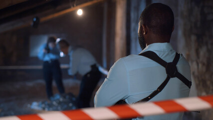 Back view of serious african officer with gun watching forensic specialists examining dead body