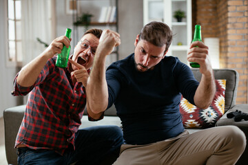 Two young friends enjoying at home. Men drinking beer and watching sports game on tv..