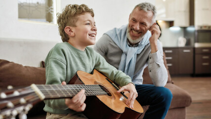 Cute little boy playing guitar and smiling while spending great time with grandfather at home, sitting together on sofa in the living room