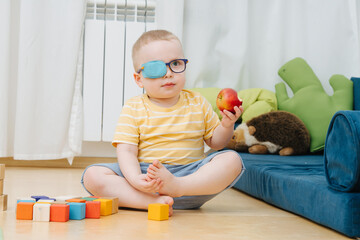 Little toddler boy in lazy eye patch eating apple on the children room