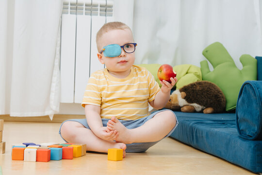 Little toddler boy in lazy eye patch eating apple on the children room