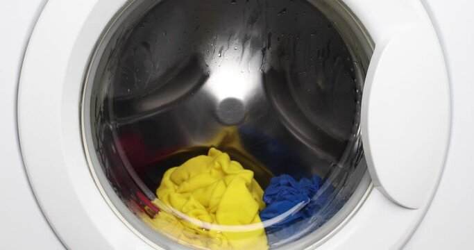 Close-up white washing machine washes multicolored T-shirts, red, yellow and blue.