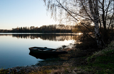 Wooden fishing boat near the river bank, the low evening sun illuminates the quiet mirror water. Trees and the sun reflected in the water. Ild River, village of Propast, Yaroslavl region, Russia.