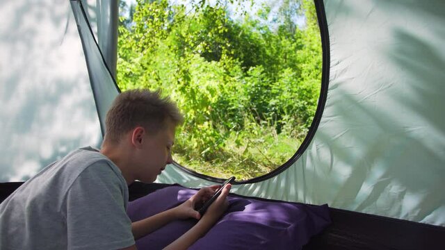 Young white kid using his mobile smartphone while laying inside of blue camping tent in summer green forest