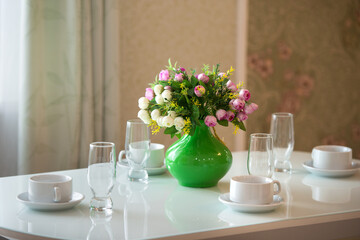 Hotel room with a white table, chairs and dishes. A green vase with artificial flowers stands on a...