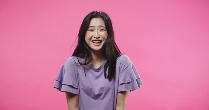 Close up portrait of cheerful beautiful young Asian woman in purple blouse standing isolated on pink background in studio talking and laughing in good mood looking at camera. Emotions concept