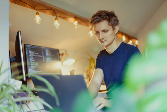 A man sitting at a desk working from home on his computer. IT specialist programmer writing code on his pc and laptop. Developer remote home office. Freelance workspace. Soft selective focus