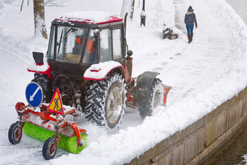 Red tractor with scoop and automated brush removes snow on embankment in park. Tractor with...