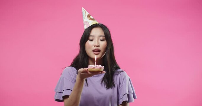 Close up portrait of joyful Asian pretty young woman in purple blouse in b-day hat blowing candle on birthday biscuit mini cake while standing on pink background celebrating making a wish