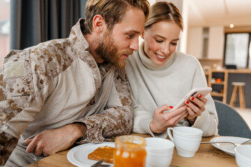 Beautiful joyful young couple using mobile phone while having breakfast