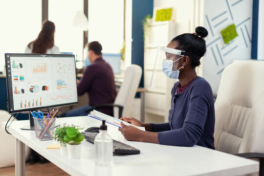 African businesswoman checking financial analysis on computer holding clipboard wearin face mask during coronavirus. Multiethnic coworkers working respecting social distance in financial company. - Powered by Adobe