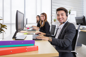 Young Asian customer service support agent operator team working in call center office with headsets and computer. Smiling and looking at camera.