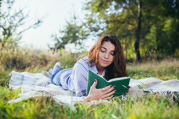 Beautiful young woman with curly black hair, resting lying on a mat in the park on the grass, and holding a book reads