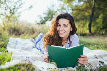 Beautiful young woman with curly black hair, resting lying on a mat in the park on the grass, and holding a book reads