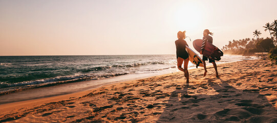 Young beautiful couple walking along the sandy beach near the ocean at sunset with surfboards, outdoor activities and sports holidays - Powered by Adobe