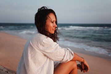Young smiling beautiful brunette woman in white shirt sitting on the beach, shot in profile