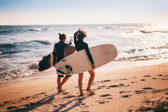 Young beautiful couple walking along the sandy beach near the ocean at sunset with surfboards, outdoor activities and sports holidays
