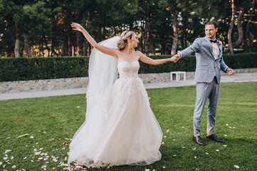 Stylish groom in a gray checkered suit and a beautiful blonde bride in a white long dress with a veil are dancing on green grass in the park in nature. Wedding portrait of newlyweds.