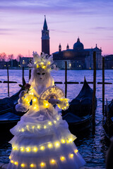 Venice, Italy - February 18, 2020: An unidentified person in a carnival costume in front of a group of gondolas and St Giorgio's Island,  attends at the Carnival of Venice.