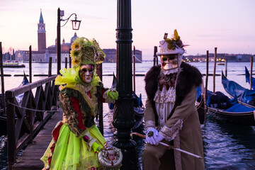 Fototapeta na wymiar Venice, Italy - February 19, 2020: An unidentified couple in a carnival costume in front of a group of gondolas and St Giorgio's Island, attends at the Carnival of Venice.