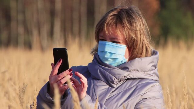 A girl in a medical mask close-up, she uses an Internet smartphone. Walk in the park in autumn. Camping on a hike. Healthy lifestyle concept. At sunset, in the evening, warm soft light.