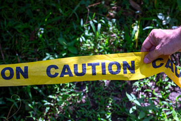 Yellow ribbon with caution inscription in man hand. Caution barricade tape on green grass. Summer park accident place surrounded by yellow caution tape. Stop sign alert ribbon