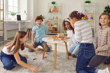 Group of excited diverse 8-10 year old children laughing at broken wood block tower while playing stacking board game together at home or in after school kids club. Fun party activities concept