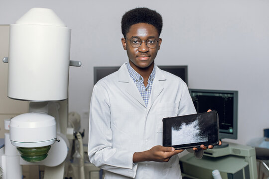 Young African American man doctor in white uniform and eyeglasses, demonstrating digital tablet pc with ultrasound scan, posing near modern ultrasonic lithotripter machine in urology medical center