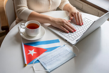 Woman tourist having breakfast with cup of coffee working on laptop.  Flag of Cuba, medical protective face mask on table of cafe. Protection from bacteria and viruse in a public place. Concept.