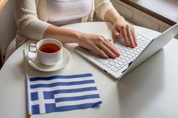 Lonely woman freelancer with flag of Greece enjoying having breakfast with cup of coffee working on laptop sitting near window in cafe at morning.