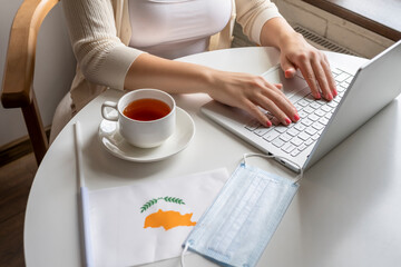 Woman tourist having breakfast with cup of coffee working on laptop.  Flag of Cyprus, medical protective face mask on table of cafe. Protection from bacteria and virus in a public place. Concept.
