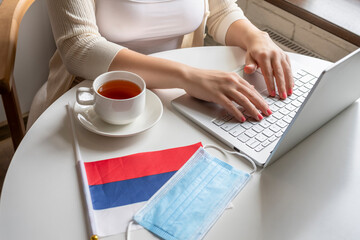 Woman tourist having breakfast with cup of coffee working on laptop.  Flag of Russia, medical protective face mask on table of cafe. Protection from bacteria and virus in a public place. Concept.