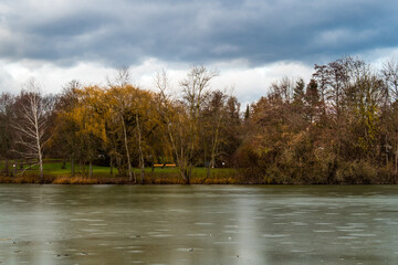 Fototapeta premium Winter landscape at frozen lake, trees reflected in the ice, dark sky in background