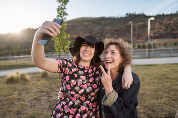 A Latina mother and daughter take a selfie photo with their cell phone where their daughter laughs and the mother makes a symbol of love and peace with her hands