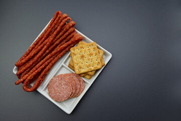 Thin boiled smoked cabanos sausage, salami slices and bran crackers in a square plate on a gray background.