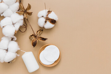 Cotton pads for removing makeup, candles and branch of cotton plant on beige background, flat lay. Spa concept. Free space for text.