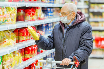Old senior european man wearing protective facial mask looking at groceries in the supermarket. Shopping during COVID-19 concept.