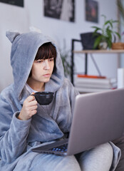 Business woman in bathrobe drinks morning coffee and surfing internet on laptop. Cheerful girl writing email on computer or working in home office. Remote studying or working concept