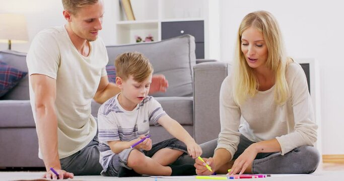 Cute little boy sitting on floor engaged in family activity paint in album together with young parents, happy loving mom and dad relax spend time with little son draw picture with coloring markers.