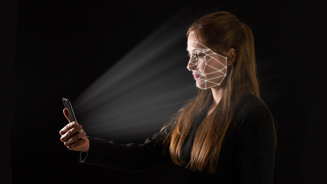 Young Caucasian businesswoman using biometric technology in her phone with light glowing on her. Concept of face recognition and identification to authorize access. Adult on dark background.