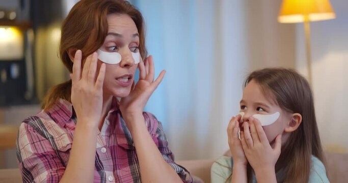 Joyful mom and daughter having beauty day at home sitting on couch and covering eyes with patches