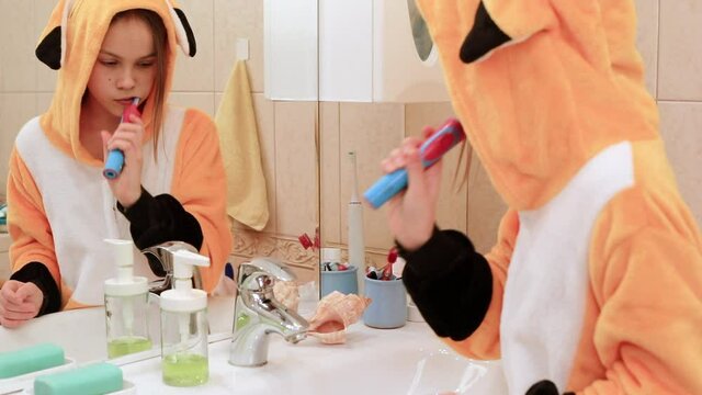 Tween girl wearing orange pyjamas brushes her teeth with electric toothbrush in a bathroom
