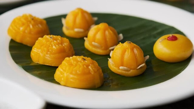 A woman's hand is delicately placing Thong-Eek, Sane Chan and Kanom Jamongkut (Thai sweets) on a white plate with banana leaves.