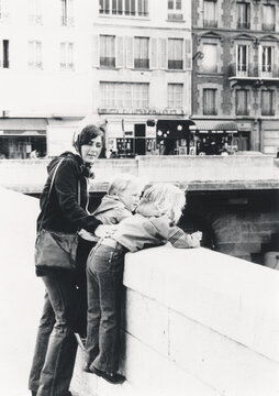 Vintage retro 1978 monochrome image of a young mother in Paris with two children hanging over bridge railing over the Seine river,  Paris, France.