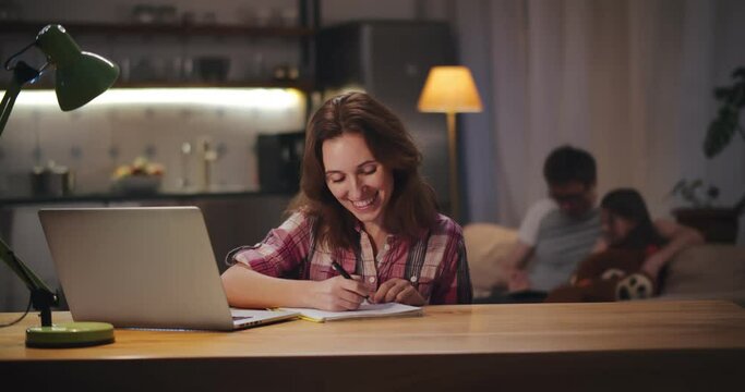 Woman having video call on laptop studying online with family relaxing on background