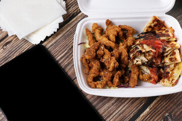 Top view of table full of food in plastic containers and black tablet. Food delivery. Chicken salad, chicken wings with sous, burger on french fries on a wooden table. Online order.