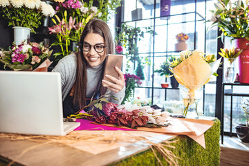 Female florist standing at her flower shop counter using mobile phone and laptop to take orders for her store. Woman flower shop owner taking online orders.