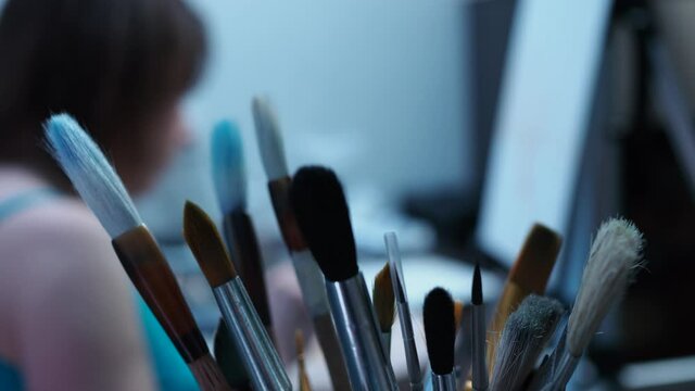 a talented artist draws with a brush on canvas. view from behind the tassels. brushes in focus, girl out of focus.