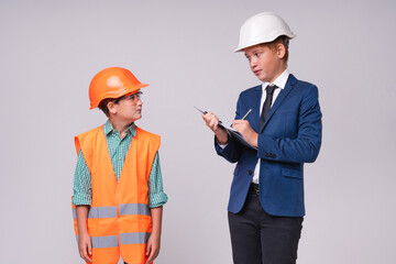 Two little caucasian boys in roles of engineer and builder in hardhats isolated over grey background
