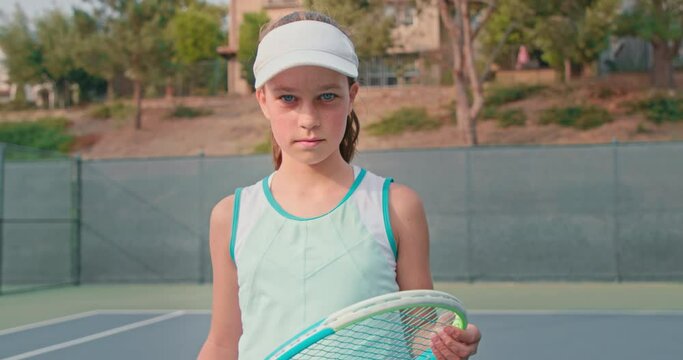 Young tennis player looks into the camera while holding her colorful racket. Girl wearing white visor and turquoise dress during the training. High quality 4k footage